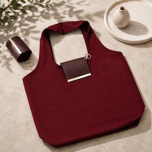 May include: A burgundy tote bag with a matching small wallet attached by a gold ring. A cylindrical case, also burgundy with a gold stripe, sits to the left. A small, white vase sits on a white plate in the background.