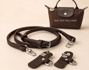 Può includere: Una tracolla per borsa in pelle marrone scuro con fibbia regolabile e moschettoni in metallo color argento, accompagnata da due linguette di fissaggio in pelle abbinate con anelli a D argentati. Una piccola borsa sullo sfondo reca la scritta BAG NOT INCLUDED.