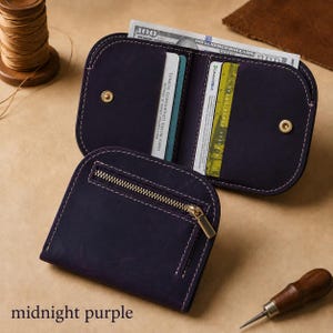 May include: A midnight purple leather wallet featuring a zippered exterior pocket, white contrast stitching, and an open interior with card slots and a currency compartment holding a one hundred dollar bill and various identification cards.