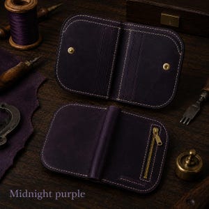 May include: A dark purple leather wallet featuring rounded corners and white stitching. The interior displays card slots and gold-tone snap buttons, while the exterior includes a vertical gold-tone metal zipper. The text Midnight purple is visible.