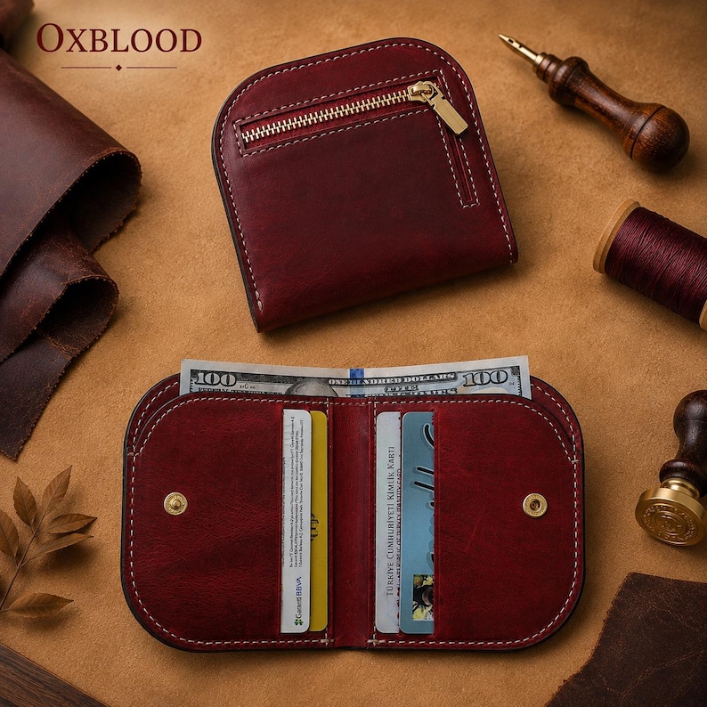 May include: An oxblood leather wallet featuring a rounded top edge, a gold-tone metal zipper pocket on the exterior, and an open interior with multiple card slots and a bill compartment containing a one hundred dollar bill with white stitching.