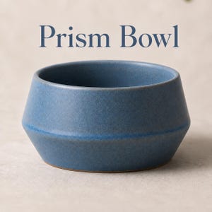 May include: A blue ceramic bowl with a matte finish and a faceted, geometric shape. The text 'Prism Bowl' is printed in dark blue letters above the bowl. The bowl features a wide base that tapers upward to a circular rim with a smooth texture.