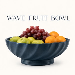 Pode incluir: Uma fruteira azul marinho com design de ondas, cheia de maçãs verdes, uvas vermelhas e laranjas. A tigela tem um design texturizado e rodopiante e uma base de pedestal. O texto "WAVE FRUIT BOWL" é exibido acima.