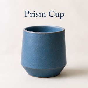 Pode incluir: Uma chávena de cerâmica azul com um acabamento mate e salpicado e uma base cónica. A chávena apresenta uma abertura larga e um rebordo arredondado e liso. O texto "Prism Cup" está impresso em letras azul-escuras acima da chávena.