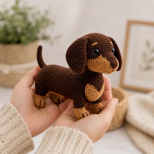 Op de afbeelding: Een handgemaakte gehaakte teckel in bruin en beige. De hond heeft een lang lichaam, korte poten en floppy oren. De hond wordt in de handen van een persoon gehouden. Een schattig en knuffelbaar speelgoed.
