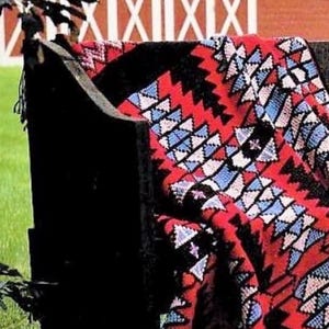 May include: A vibrant red, black, and blue patterned blanket draped over a black bench. The blanket features a geometric design with triangular shapes. The background includes a red barn with white accents and a green lawn.
