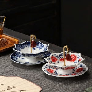 May include: Two decorative ceramic bowls with matching saucers. The bowls have a unique floral shape and are decorated with blue and white floral patterns. Each bowl has a gold-colored handle with a red tassel. The saucers also feature matching floral designs.