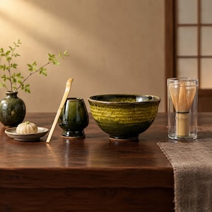 May include: A matcha tea set featuring a dark green ceramic bowl, a matching whisk holder, a bamboo scoop, a small plate with a light-coloured confection, a green vase with a leafy branch, and a bamboo whisk inside a clear cylindrical case.