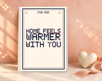May include: A rectangular greeting card with a cream background and a dark blue and red border. The text FOR MOM is at the top, and the text HOME FEELS WARMER WITH YOU is centered in a dark blue pixelated font with a subtle red shadow effect.