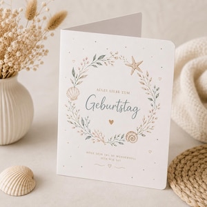 Könnte beinhalten: Eine gefaltete Geburtstagskarte mit Muschel- und Seestern-Design. Die Karte zeigt einen Kranz aus Blättern und Muscheln um den Text "Alles Liebe zum Geburtstag". Eine kleine Muschel ist im Vordergrund zu sehen.