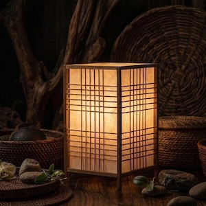 Könnte beinhalten: Eine quadratische japanische Lampe aus Holz mit einem Gittermuster. Die Lampe strahlt ein warmes, einladendes Licht aus. Sie steht auf einer Holzoberfläche, mit natürlichen Elementen im Hintergrund.