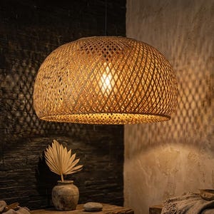 Puede incluir: Una lámpara colgante de bambú tejido. La luz en forma de cúpula tiene un diseño de celosía, proyectando sombras. La luz cálida ilumina el interior, creando un ambiente acogedor. La lámpara cuelga de un cable negro.