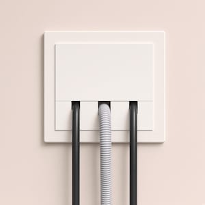 Puede incluir: Una cubierta cuadrada blanca para cables montada en la pared con tres ranuras verticales. Dos cables negros lisos están colocados en las ranuras exteriores, mientras que un cable gris estriado está centrado en la ranura central, extendiéndose hacia abajo.