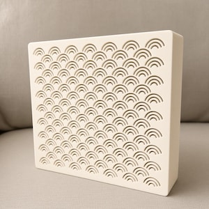 May include: A rectangular off-white block featuring a repeating pattern of recessed concentric semi-circles across its front surface. The object has a smooth, matte finish and a solid, boxy structure with clean, sharp edges and a uniform light color.