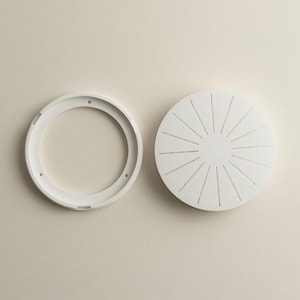 May include: A white circular ring component with three small mounting holes and a solid white circular cover featuring a radial pattern of thin, evenly spaced engraved lines extending from the center to the outer edge of the disc.