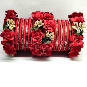 Può includere: Un set di bracciali rigidi avvolti in seta rossa con fasce metalliche dorate e grappoli di rose in tessuto rosso con piccoli accenti dorati. Il testo TheBrassHeritage JEWELRY è visibile al centro della pila di bracciali.
