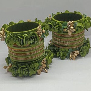 May include: Two olive green bangles featuring a stacked design wrapped in thread. Each bangle is adorned with fabric flower petals in matching green tones and small clusters of gold-colored metallic beads along the top and bottom edges.