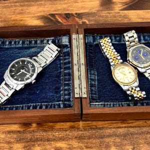 May include: A rectangular wooden display box with a hinged lid, featuring dark blue denim fabric lining the interior. Three wristwatches are stored inside: one with a black dial and silver band, and two with metallic bands and gold accents.