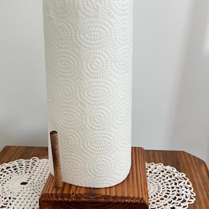 May include: A paper towel holder featuring a rectangular dark brown stained wooden base with a vertical wooden dowel and a central upright post. A roll of white paper towels with a textured spiral pattern is mounted on the central post.