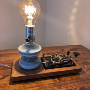 May include: A table lamp featuring a glowing Edison-style light bulb mounted on a white ceramic insulator, which is attached to a rectangular wooden base alongside a vintage black metal telegraph key with visible metal components and wiring.