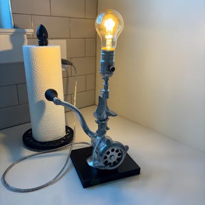 May include: A unique lamp made from a vintage meat grinder, featuring a clear light bulb. The lamp is mounted on a black square base and has a silver-colored metal finish. The light is turned on, illuminating the room.
