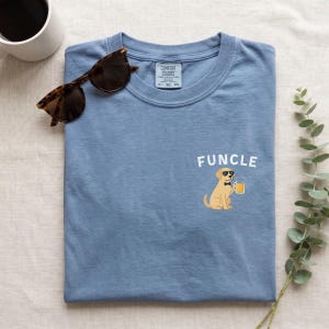 May include: A blue crew neck t-shirt featuring a graphic of a yellow dog wearing sunglasses and a bow tie, holding a glass of beer. The text "FUNCLE" is printed above the dog. The shirt has a Comfort Colors brand label on the inner neckline.