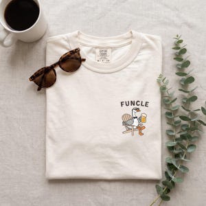May include: A cream-colored t-shirt featuring a graphic of a goose wearing sunglasses and a bowtie, sitting in a wooden chair while holding a beer mug. The text "FUNCLE" is printed above the goose. The shirt has a Comfort Colors brand label.