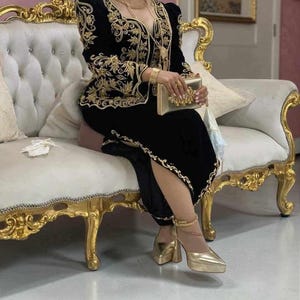 May include: A black velvet two-piece outfit featuring intricate gold embroidery along the neckline, bodice, and hem. The ensemble includes a long-sleeved top and a matching skirt, paired with a gold clutch and metallic gold pointed-toe platform heels.