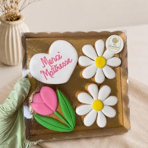 Peut inclure: Une boîte cadeau carrée contenant quatre biscuits décorés : un cœur blanc avec le texte rose Merci Maîtresse, deux marguerites blanches à cœur jaune et une tulipe rose avec des feuilles vertes. Les biscuits sont disposés sur un fond doré.