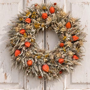 May include: A circular wreath composed of dried wheat stalks, grasses, and small yellow flowers, accented with bright orange physalis husks. The arrangement features a mix of beige, tan, and muted green tones, creating a textured, natural appearance.