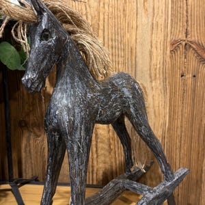 Puede incluir: Un caballo mecedora de madera en color gris carbón con un acabado desgastado que revela tonos de madera más claros. El caballo presenta una crin y una cola texturizadas hechas de cuerda de yute color canela, sobre dos balancines.