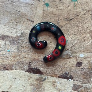 May include: A black glass spiral ear gauge featuring embedded millefiori patterns in red, blue, yellow, and white. The jewelry has a smooth, glossy finish with a curved, tapered shape that transitions from a thicker end to a thinner point.