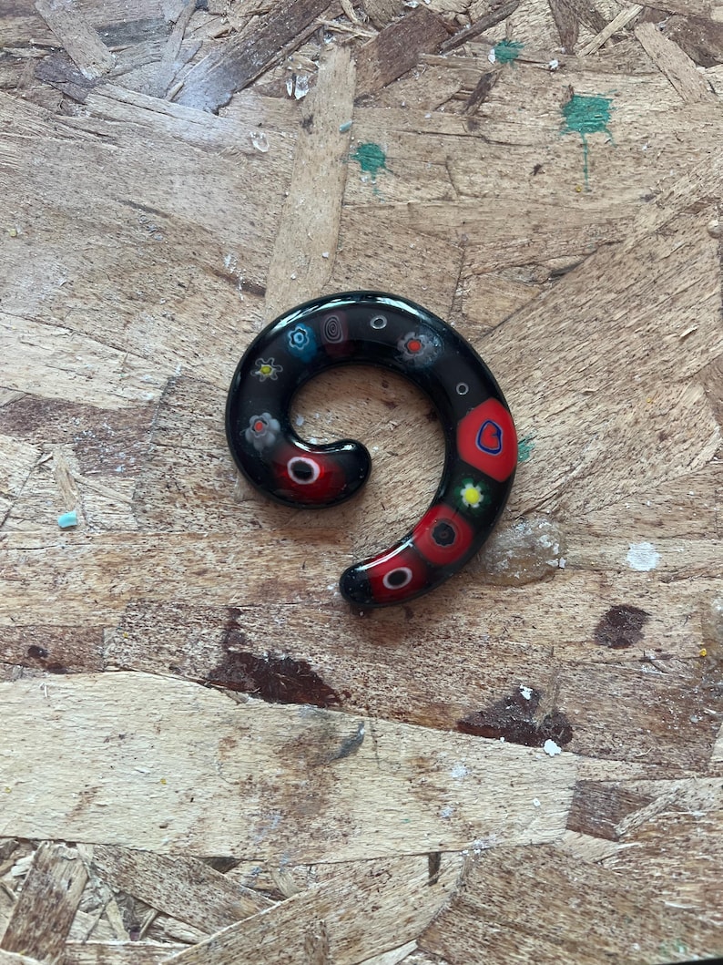 May include: A black glass spiral ear gauge featuring embedded millefiori patterns in red, blue, yellow, and white. The jewelry has a smooth, glossy finish with a curved, tapered shape that transitions from a thicker end to a thinner point.