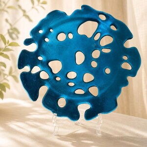 Op de afbeelding: Een cirkelvormig, metallic blauw decoratief bord met een onregelmatige, organische rand en talloze uitgesneden gaten van verschillende groottes over het hele oppervlak. Het bord rust op een doorzichtige kunststof displaystandaard.