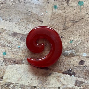 May include: A deep red, translucent spiral-shaped ear gauge plug. The jewelry features a smooth, polished surface with a curved, tapered design that coils inward toward the center, creating a distinct circular silhouette for body modification.