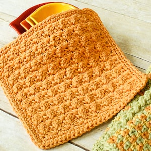 May include: A crocheted pot holder in a warm orange color. The pot holder is made with a textured stitch that creates a raised pattern.