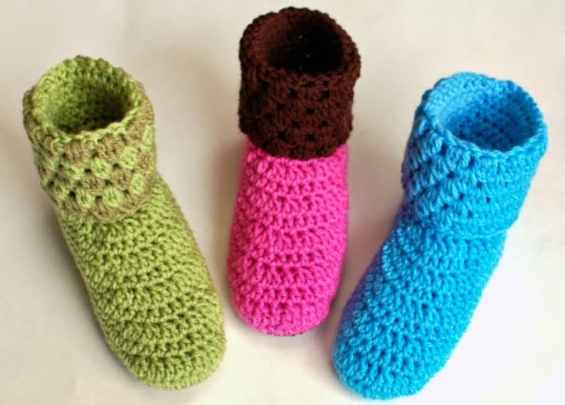 May include: Three pairs of crocheted slippers in green, pink, and blue. The slippers have a fold-over cuff and are made with a simple stitch pattern.