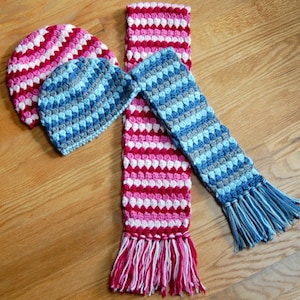 May include: Two crocheted hats and a scarf in pink, white, blue, and gray. The hats are striped and the scarf has a bobble stitch pattern.