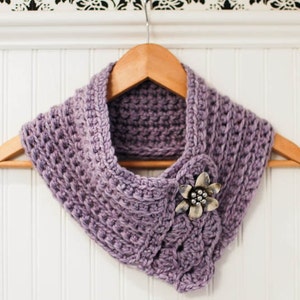 May include: A purple crocheted cowl with a silver flower brooch. The cowl is hanging on a wooden hanger.