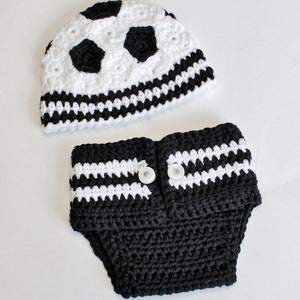 May include: A handmade crochet baby set featuring a soccer ball design. The set includes a white and black hat with soccer ball detailing and a matching diaper cover with white stripes and two white buttons. The set is made of soft yarn.