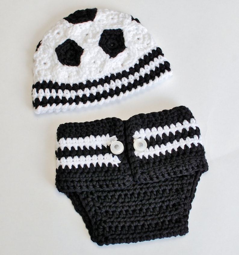 May include: A handmade crochet baby set featuring a soccer ball design. The set includes a white and black hat with soccer ball detailing and a matching diaper cover with white stripes and two white buttons. The set is made of soft yarn.