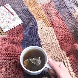 Könnte beinhalten: Eine gehäkelte Decke in verschiedenen Farben, darunter Braun, Beige, Lila und Rosa. Eine Tasse Tee mit einem Teebeutel steht auf der Decke, und ein Notizbuch mit Blumenmuster liegt auf der Decke.