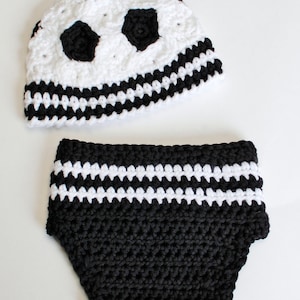 May include: A handmade crochet baby set in black and white. The set includes a hat with soccer ball design and matching diaper cover. The hat features black pentagons on a white background, and the diaper cover has white stripes.