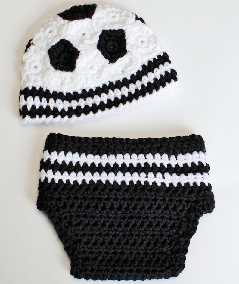 May include: A handmade crochet baby set in black and white. The set includes a hat with soccer ball design and matching diaper cover. The hat features black pentagons on a white background, and the diaper cover has white stripes.