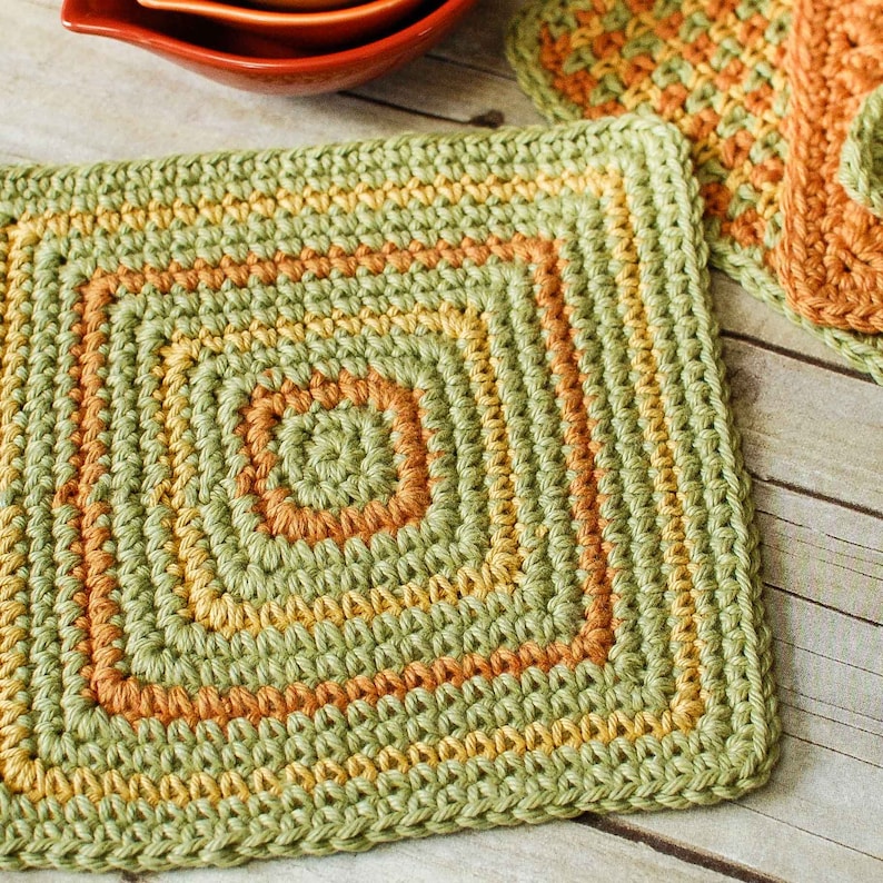 May include: Green, yellow, and orange crocheted square pot holder. The pot holder has a square shape with a smaller square in the center.