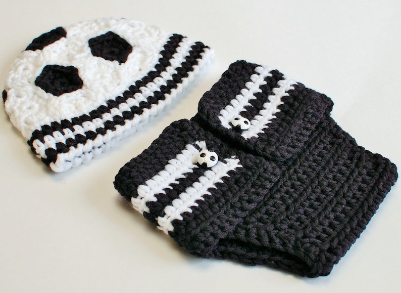 May include: A crocheted soccer-themed baby set, featuring a white and black soccer ball hat and matching leg warmers. The leg warmers are dark gray with white stripes and soccer ball buttons.