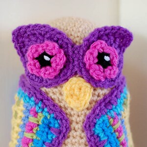 May include: A handmade crochet owl with a beige body, purple wings, and pink flower accents. The owl has a yellow beak and a colorful vest with blue, green, and pink squares. The eyes are black with white highlights.