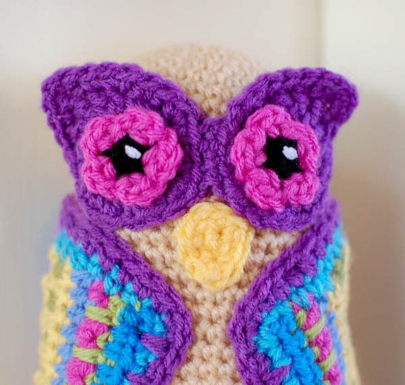 May include: A handmade crochet owl with a beige body, purple wings, and pink flower accents. The owl has a yellow beak and a colorful vest with blue, green, and pink squares. The eyes are black with white highlights.