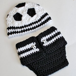 May include: A crocheted baby set featuring a soccer ball design. The set includes a white and black hat with soccer ball patches and a matching diaper cover. The diaper cover has white stripes and soccer ball buttons.