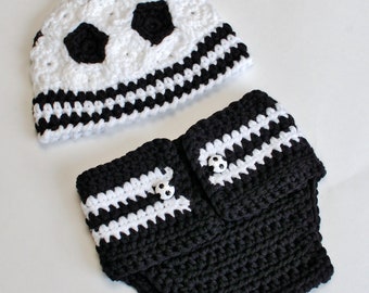May include: A crocheted baby set featuring a soccer ball design. The set includes a white and black hat with soccer ball patches and a matching diaper cover. The diaper cover has white stripes and soccer ball buttons.